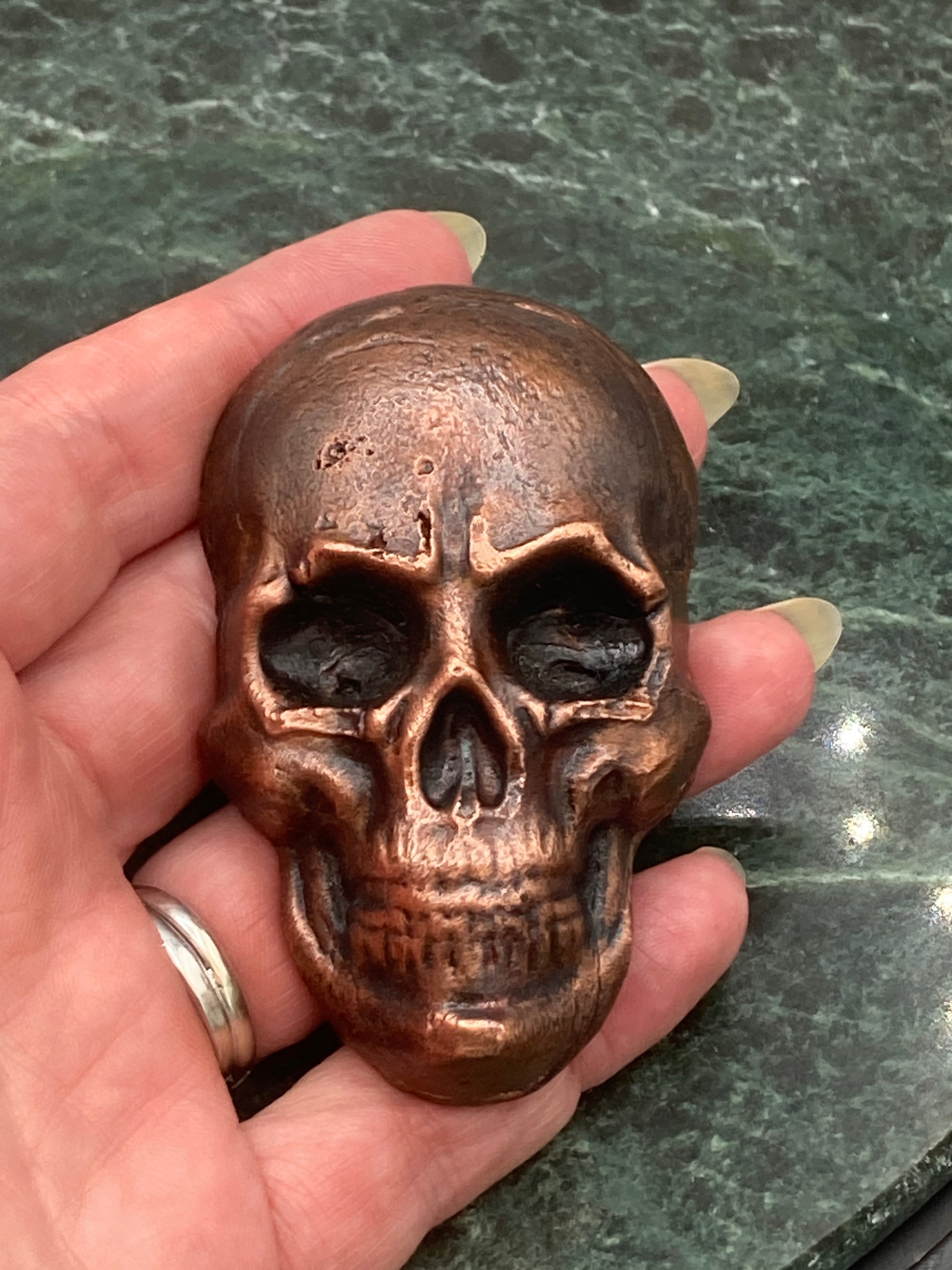 Bronze skull-shaped object held in a hand against a green marble background