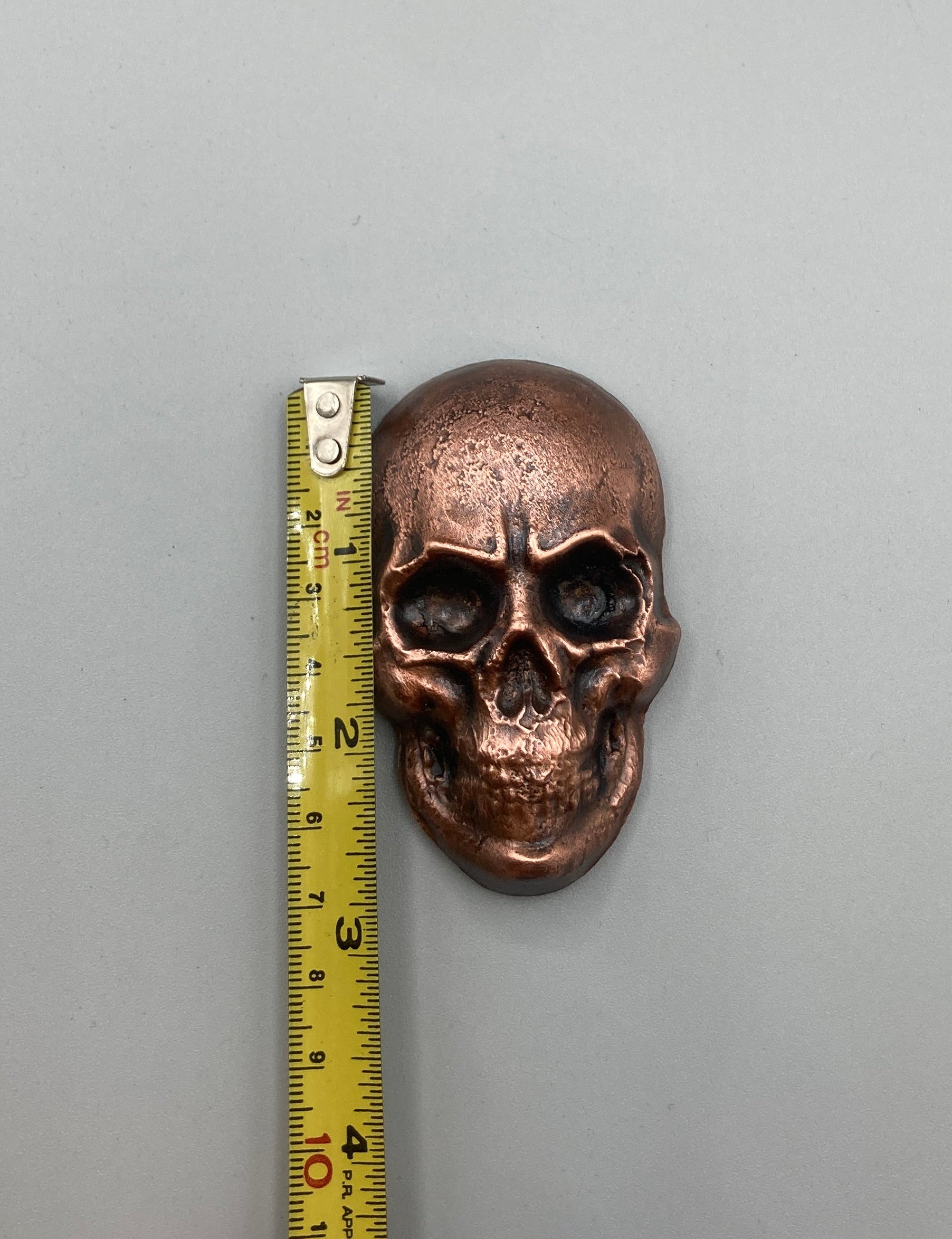 Bronze skull-shaped object with a measuring tape for scale on a gray background